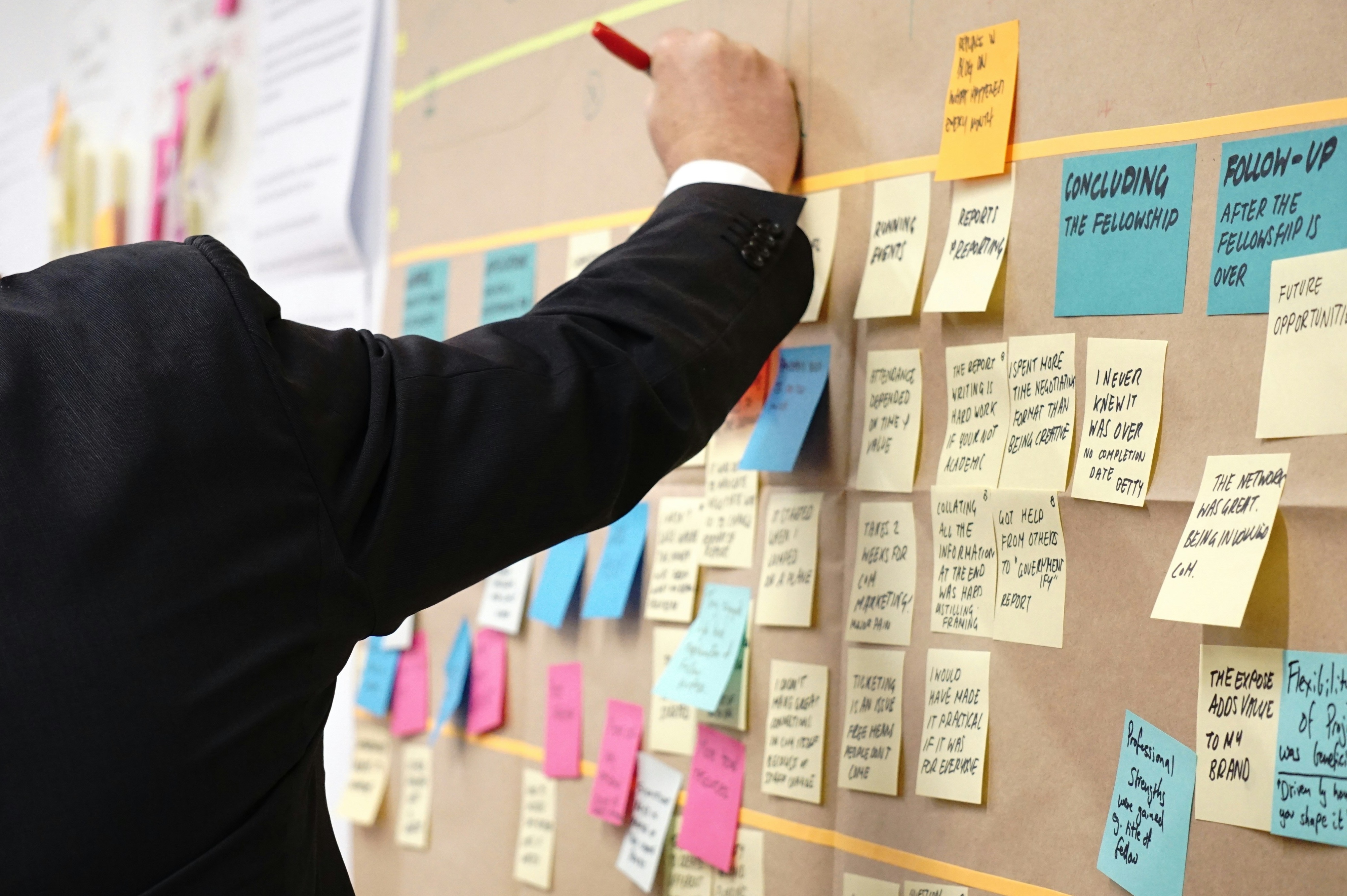 A suited individual writing on a post it on a large, filled corkboard.