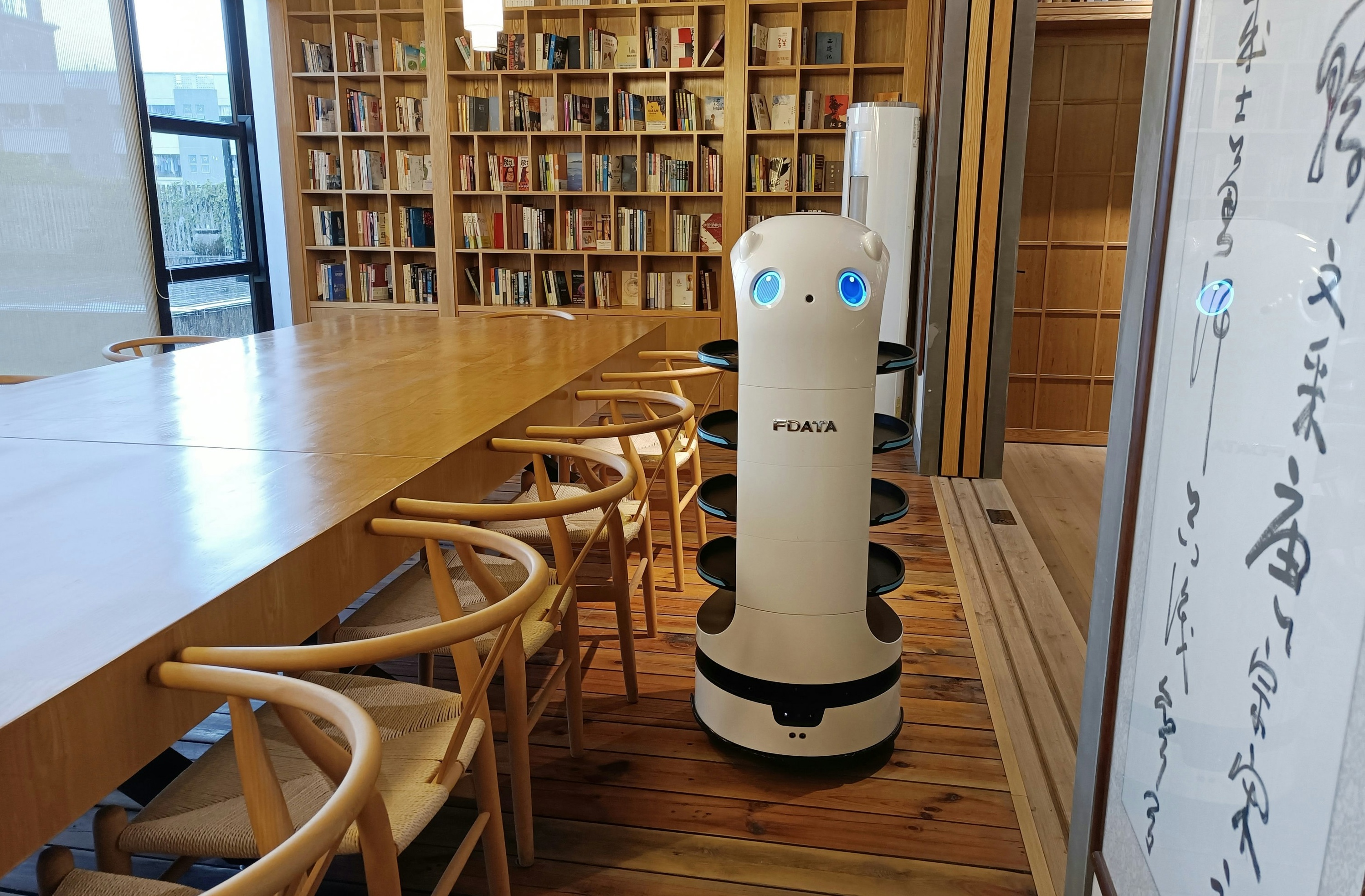 Some kind of robot, white and table height, with blue light eyes in a small library or study room.